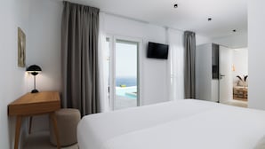 A modern hotel room with a large bed, a desk, a chair, a television, and a view of the ocean through a sliding door.