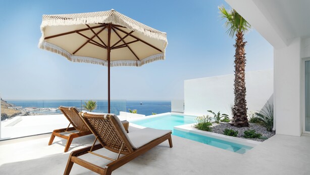 A poolside area with two wooden lounge chairs and a large umbrella, overlooking a clear blue sea and a modern building.