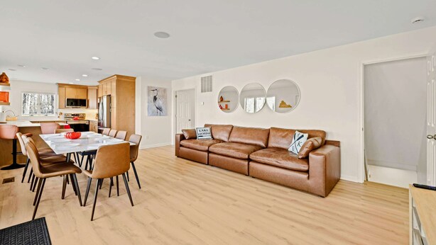 A modern living room with a brown leather sofa, a dining table with chairs, and a kitchen in the background. - Lakefront Lake Anna with Dock, Hot Tub, Game Room (Bumpass)