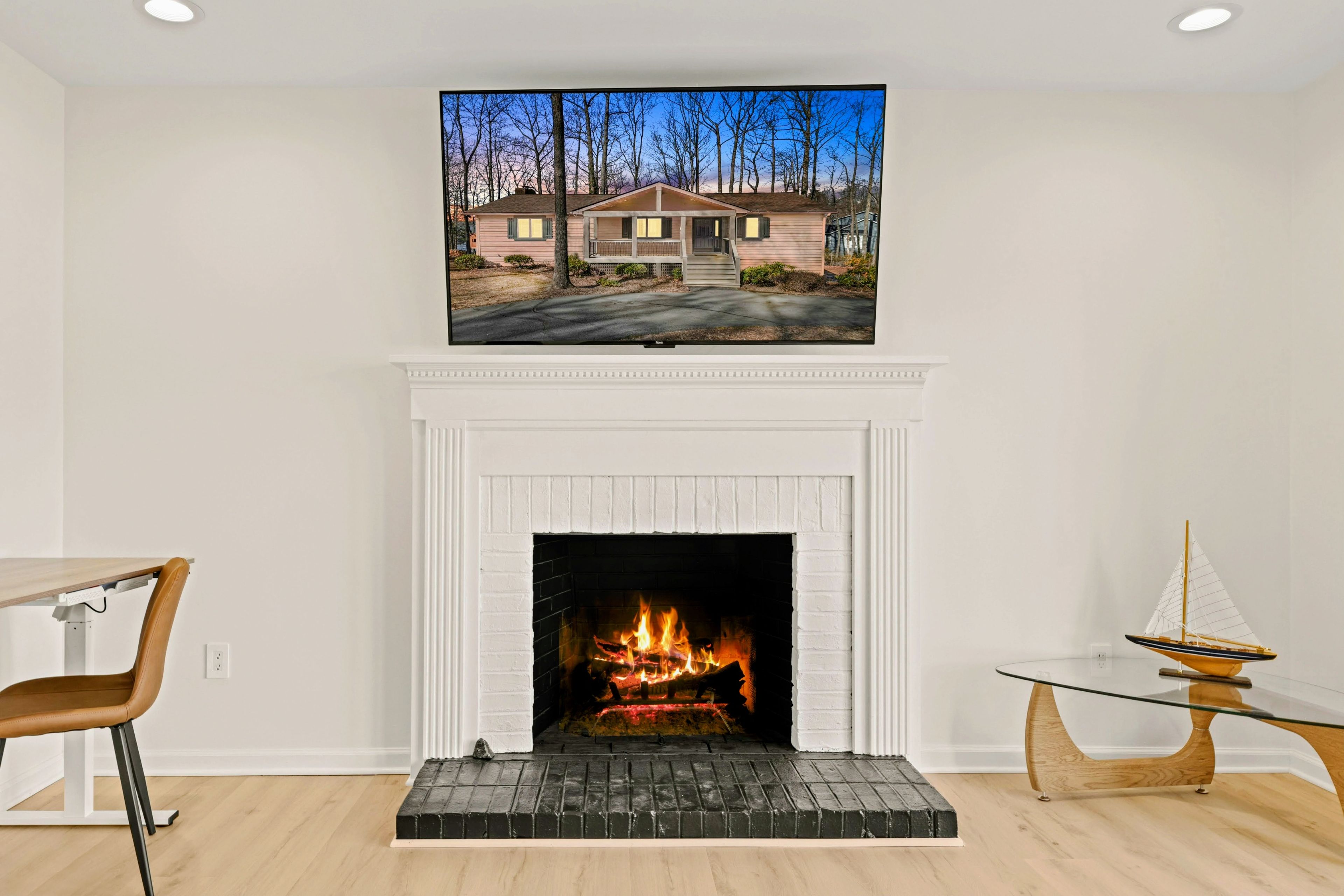 Una habitación con chimenea, una mesa de comedor con sillas y un televisor sobre la chimenea mostrando una casa.