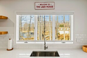 Una cocina con un lavamanos de acero inoxidable, una ventana con persianas y un letrero sobre la ventana que dice 'WELCOME TO THE LAKE HOUSE'.