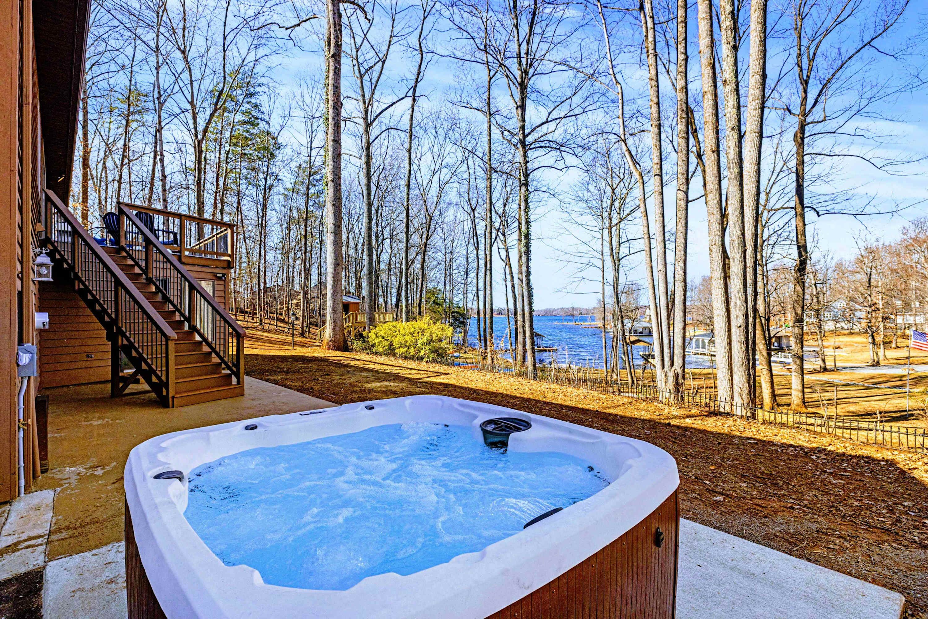 A hot tub with a wooden deck and stairs leading to a covered area, surrounded by trees and a view of a body of water.