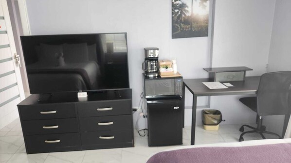 A compact hotel room with a bed, a desk, a small refrigerator, a coffee maker, and a television.