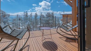 Um deck de madeira com duas cadeiras de lounge e uma pequena mesa, com vista para uma paisagem montanhosa coberta de neve.