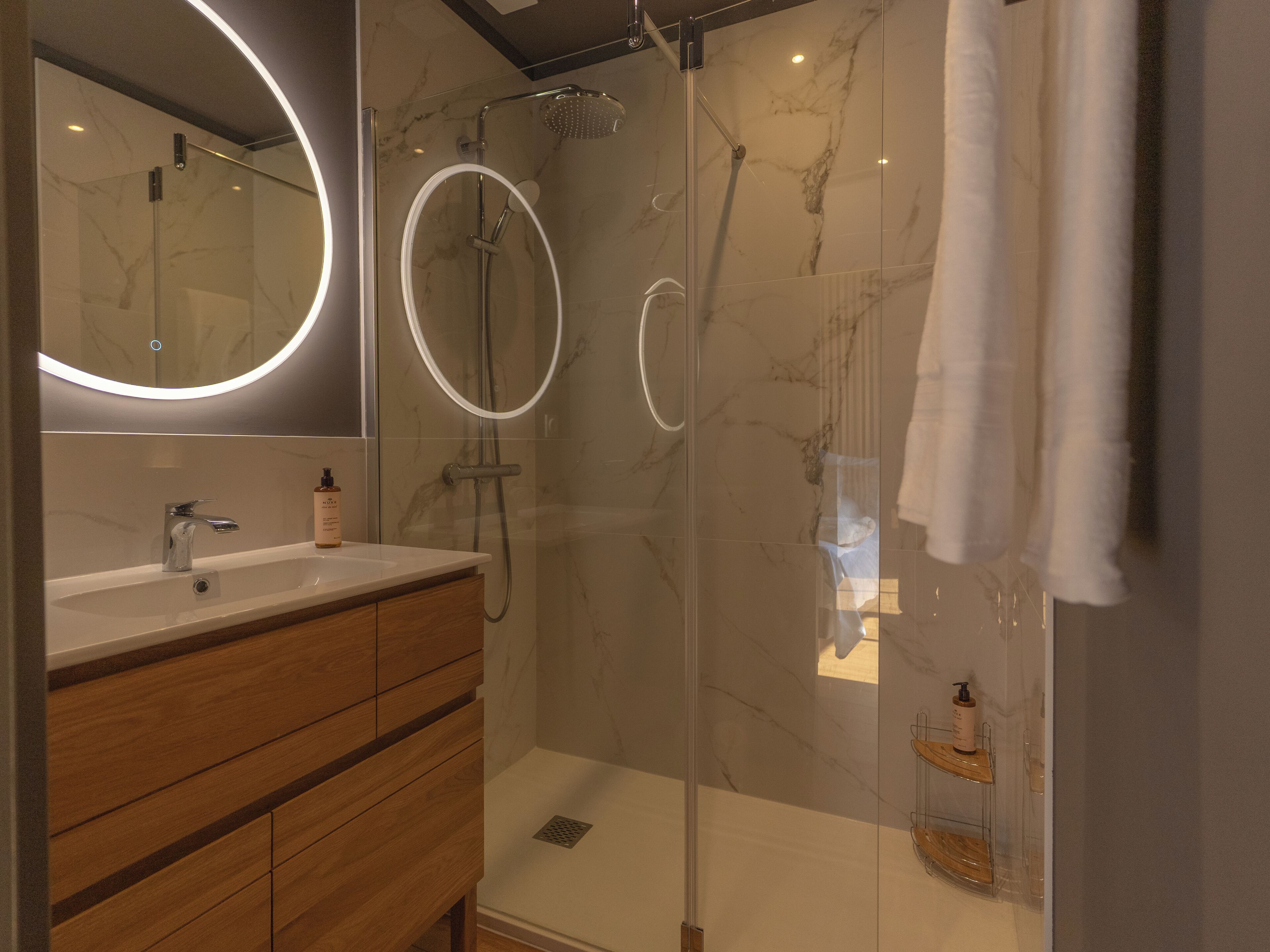 A modern bathroom with a glass-enclosed shower, a wooden vanity, and a large mirror with a circular light.