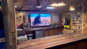A rustic bar area with a television displaying a nature scene, a 'No Contractor Parking' sign, and a Pac-Man arcade machine.