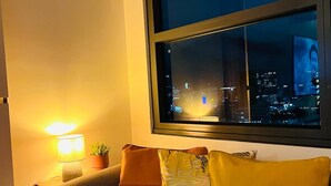 A modern living room with a sofa, cushions, a lamp, and a window overlooking a cityscape at night. - Sassy and Classy Fully Furnished Studio Unit located Cebu City Centre (Cebu City)