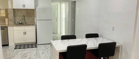 Eine moderne Küche mit weißen Schränken, einer Marmorarbeitsplatte und einem integrierten Kühlschrank. Es gibt einen Essbereich mit einem Marmortisch und schwarzen Barhocker.