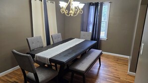 Un comedor con una mesa rectangular, seis sillas grises tapizadas, un banco, una araña y una ventana con cortinas.