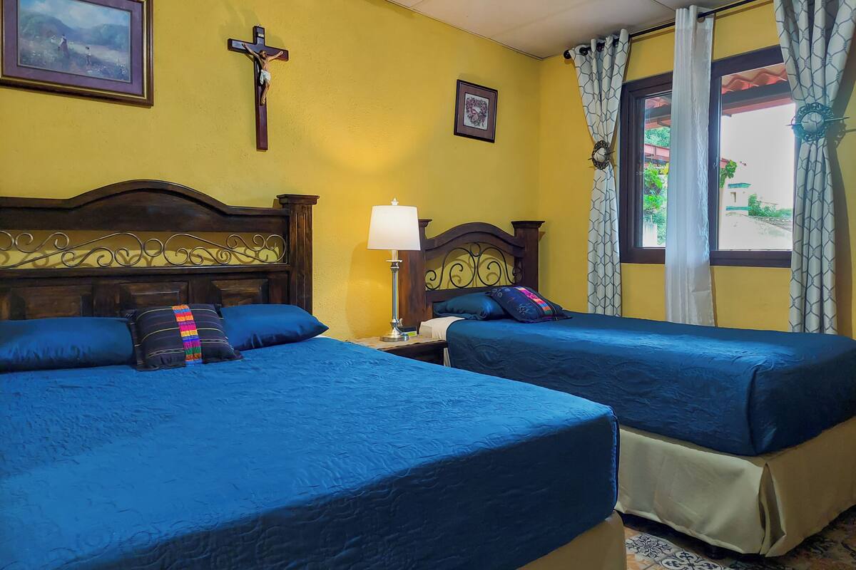 Habitación de hotel con dos camas, cabecera de madera, mesita de noche con lámpara y ventana con cortinas.