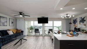 Ein modernes Wohnzimmer mit einem dunkelblauen Sofa, einem Flachbildfernseher und einem Balkon mit Blick auf ein Gewässer.