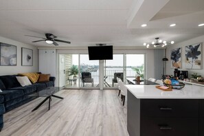 Uma sala de estar moderna com um sofá azul escuro, uma TV de tela plana e uma varanda com vista para um corpo de água.