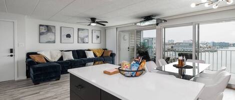 Ein modernes Wohnzimmer mit einem dunkelblauen Sofa, einem weißen Couchtisch und einem Essbereich mit weißem Tisch und Stühlen. Es gibt Deckenventilatoren, ein großes Fenster mit Blick auf die Stadt und Kunstwerke an den Wänden.
