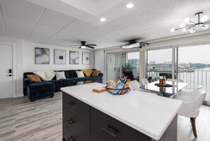 Un soggiorno moderno con un divano blu scuro, un tavolino da caffè bianco e una zona pranzo con tavolo e sedie bianche. Sono presenti delle ventole a soffitto, un'ampia finestra con vista sulla città e opere d'arte alle pareti.