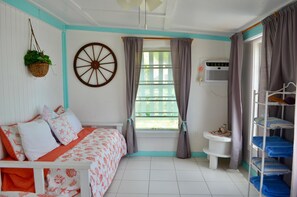 Une chambre avec un lit, une fenêtre avec des rideaux, un climatiseur fixé au mur, une petite table avec un vase et une étagère avec des serviettes pliées.