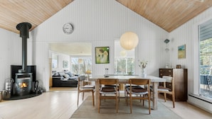 A spacious living room with a large wooden ceiling, a black wood stove, a dining area with wooden chairs, and a sofa in the background. - 3 bedroom awesome home in Ebeltoft (Ebeltoft)