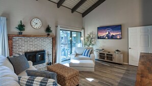 A living room with a fireplace, a sofa, a coffee table, and a television. There is a door and a window in the background.