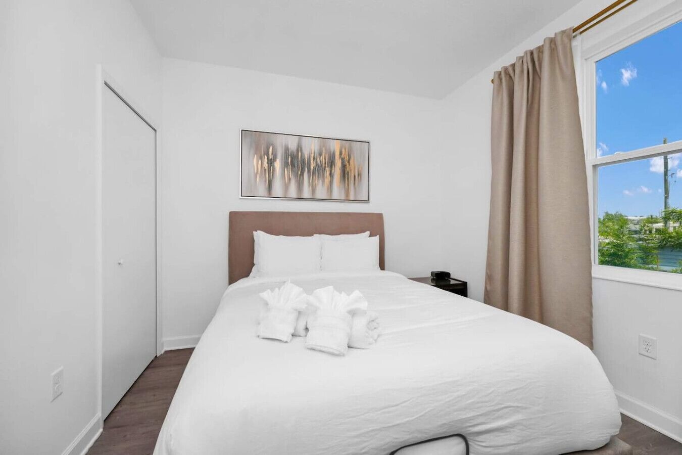 Une chambre d’hôtel avec un grand lit, une table de chevet, un tableau au-dessus du lit et une fenêtre avec des rideaux.
