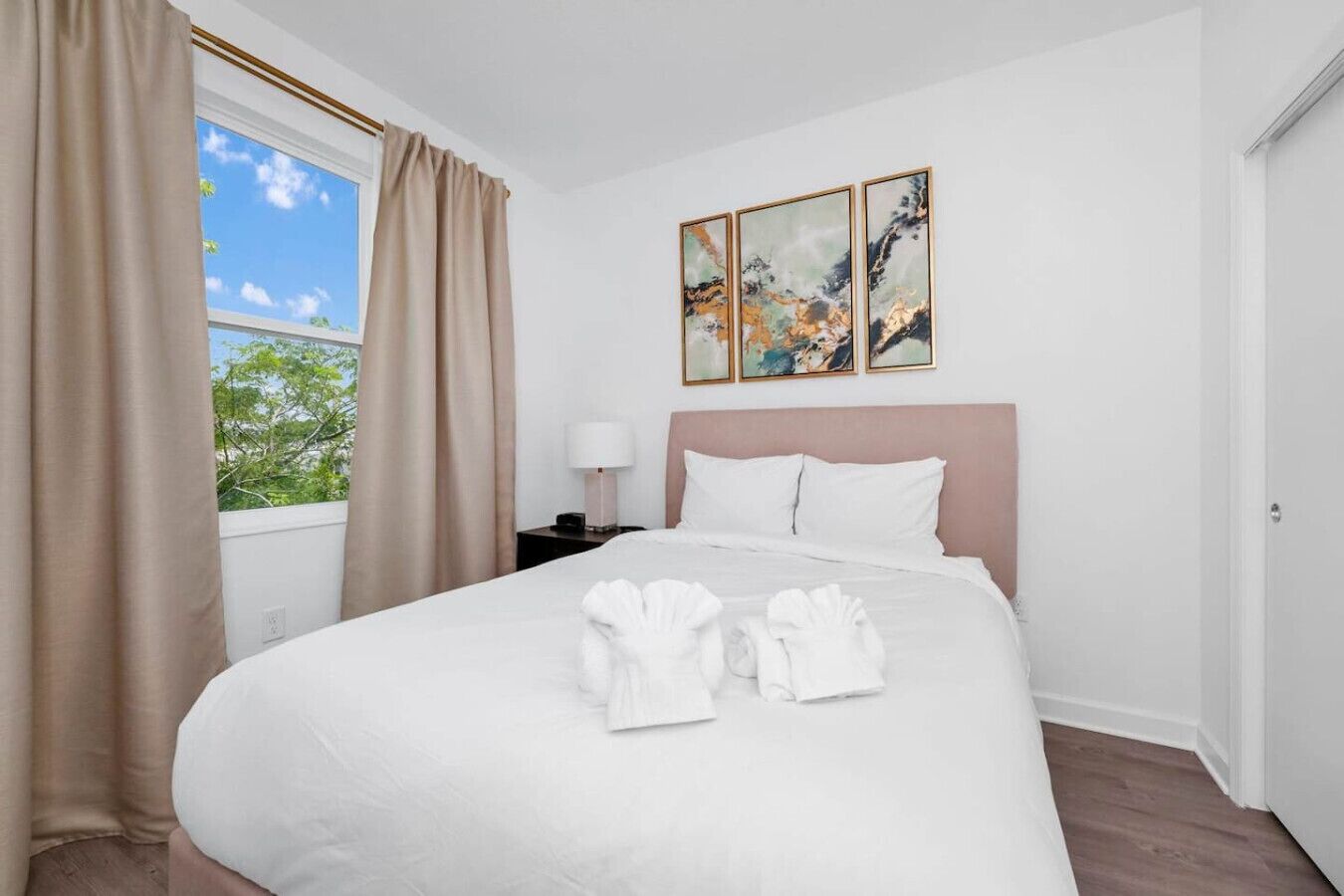 Ein Hotelzimmer mit einem großen Bett, beigen Vorhängen, einem Nachttisch mit Lampe und drei gerahmten Kunstwerken an der Wand.