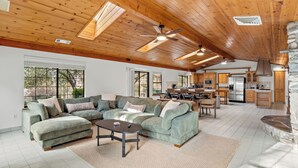 A spacious living room with a sectional sofa, a coffee table, and a dining area with chairs. A kitchen with wooden cabinets and a stone accent wall is visible in the background.