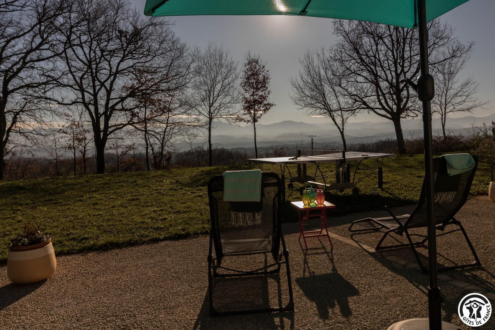 Ein Sitzbereich im Freien mit Tisch und Stühlen, einem Blumentopf und Blick auf Bäume und Berge im Hintergrund.