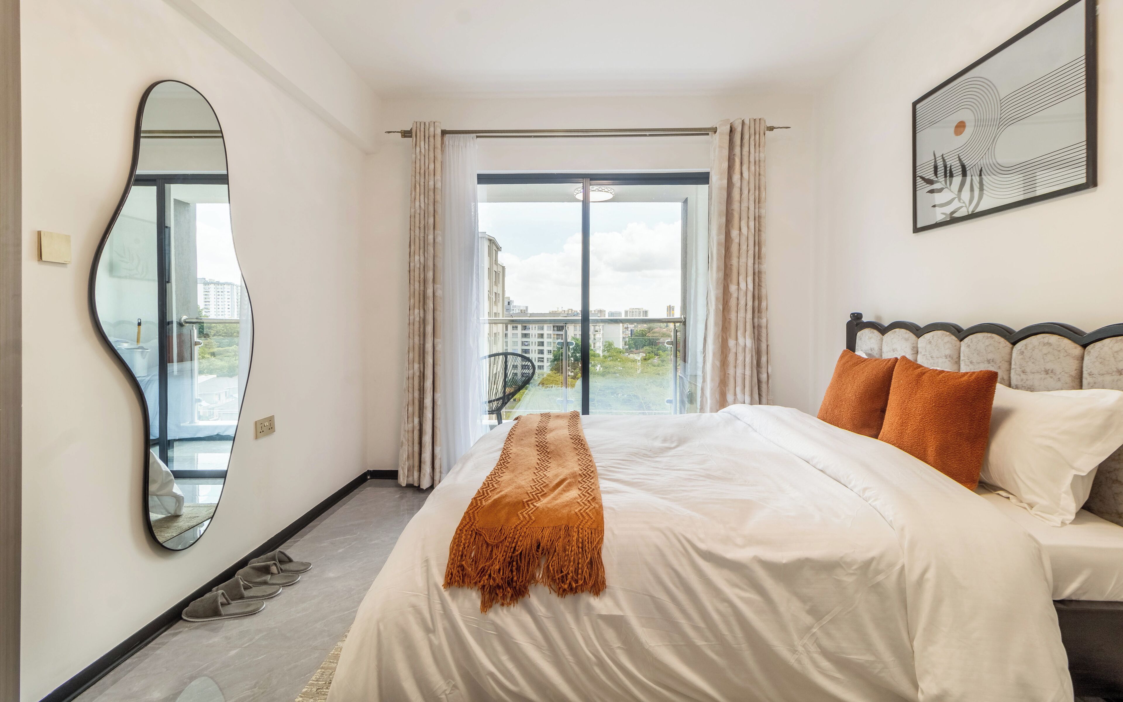 A hotel room with a large bed, a view of the city through a balcony, and a modern mirror.