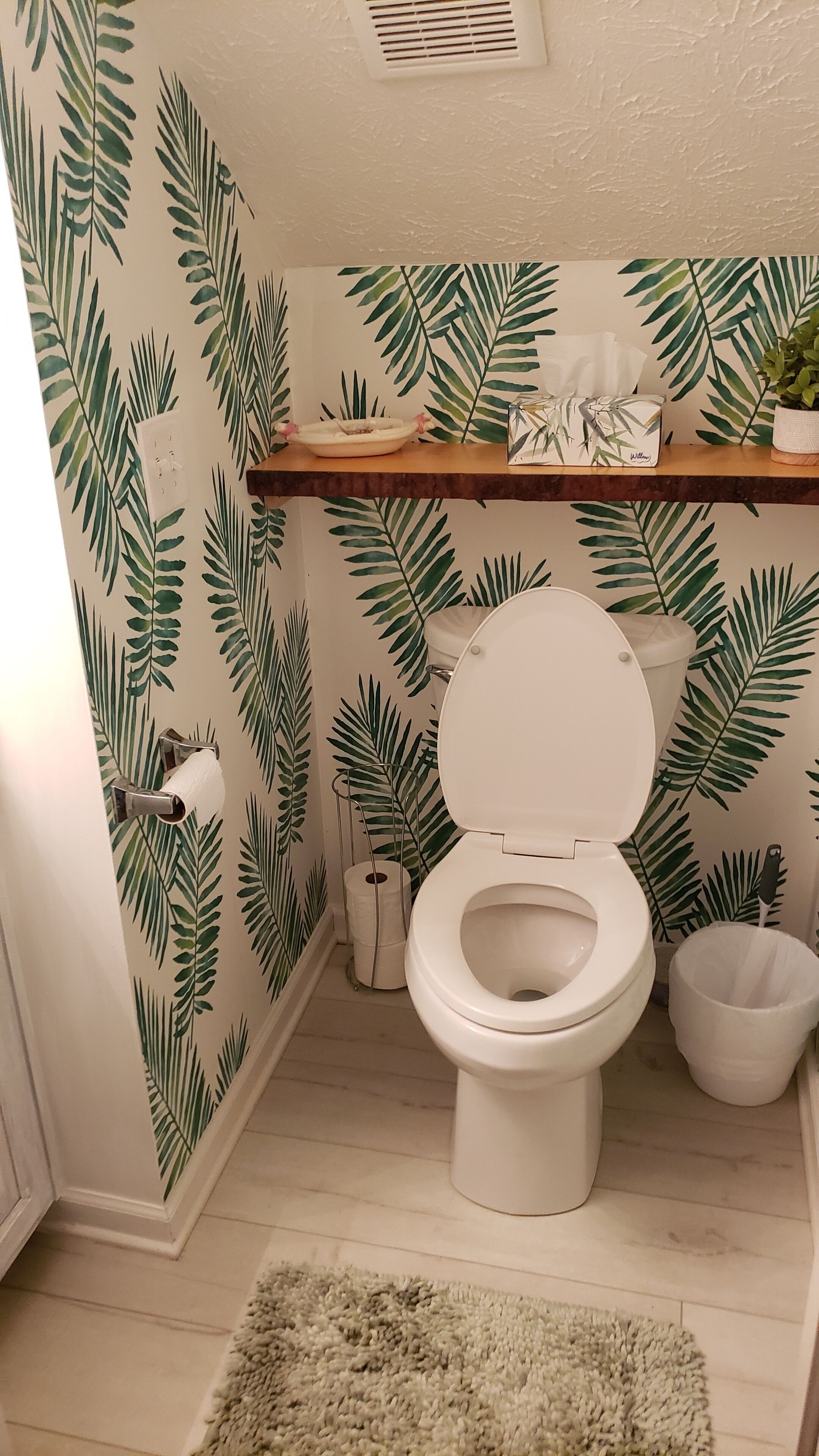 Un bagno con il water, uno scaffale con articoli da toilette, una pianta e una parete decorata con un motivo a foglia.