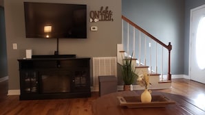 A living room with a flat-screen TV, a dark wooden entertainment center, a staircase, and a small table with a vase.