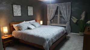 A bedroom with a wooden bed, two bedside lamps, a window with curtains, and two framed floral wall art pieces.