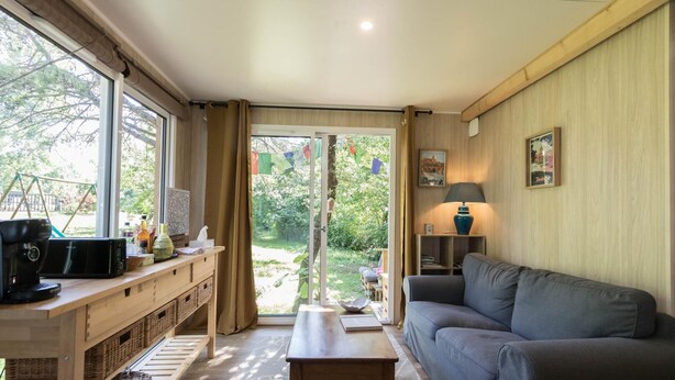 A cozy living room with a sofa, a coffee table, and a wooden side table with a lamp. A large window offers a view of greenery outside. - Les Collines du Pastel (VERFEIL)