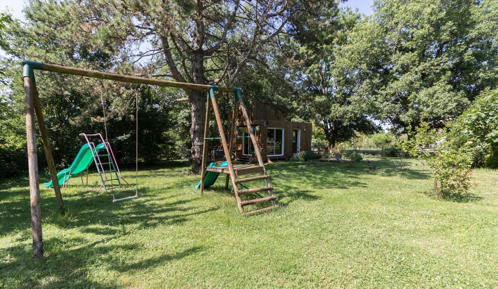 Ein Spielplatz mit Schaukeln und einer Rutsche, umgeben von Bäumen und einem kleinen Gebäude.