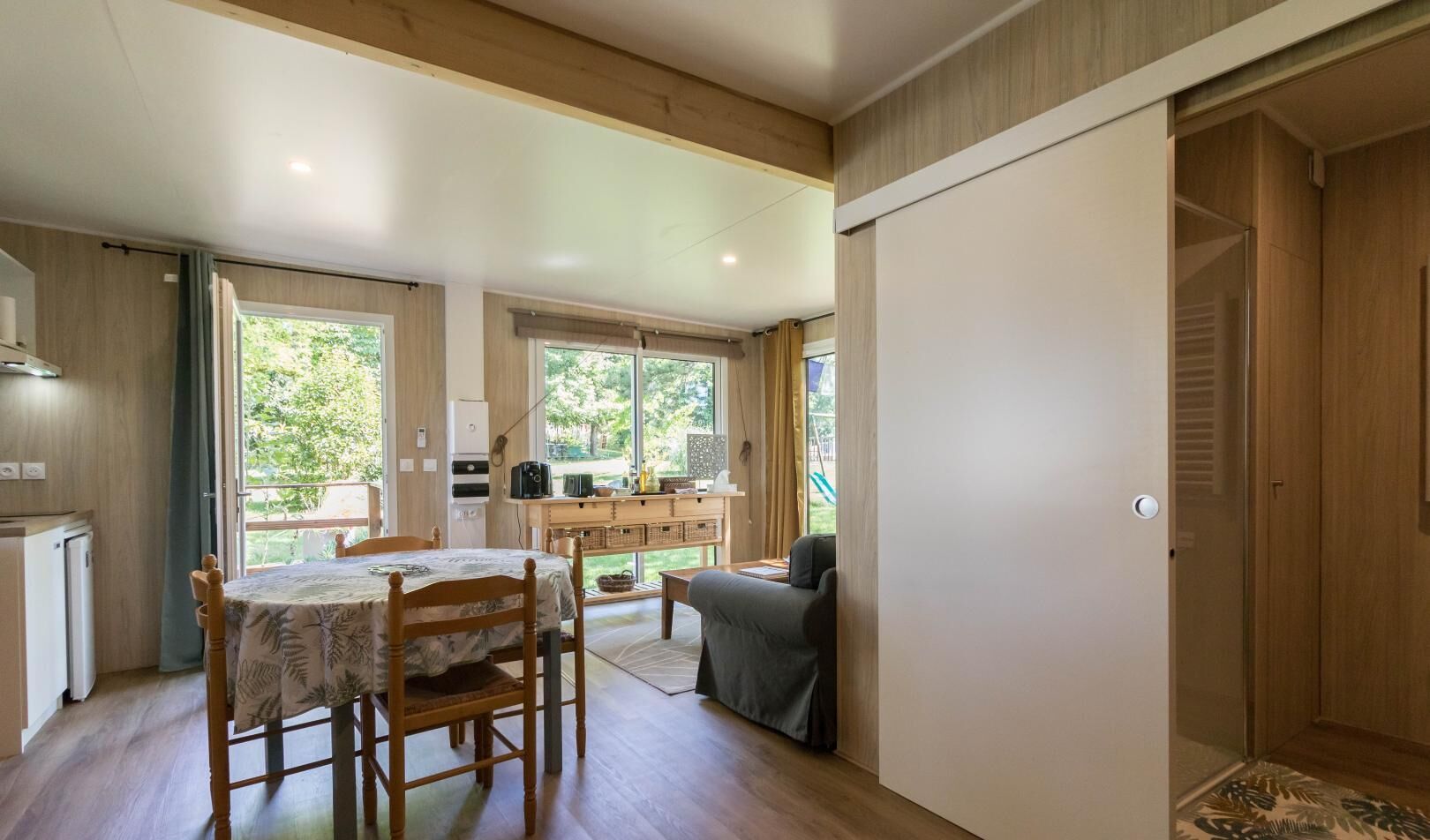 Une pièce spacieuse comprenant une table et des chaises, un canapé, un coin cuisine et de grandes fenêtres donnant sur l’extérieur.