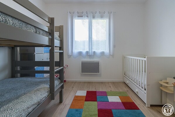 A child's bedroom with bunk beds, a radiator, a window with curtains, and a colorful rug. - A l'Orée du Roc - FRONTON (FRONTON)