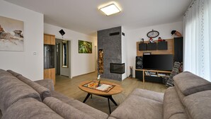 Una sala de estar moderna con un sofá gris, una mesa de centro con camino de mesa floral, un televisor de pantalla plana, una chimenea y un reloj en la pared.