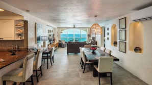 A spacious dining area with a view of the ocean, a kitchen with a tiled backsplash, and a ceiling fan.