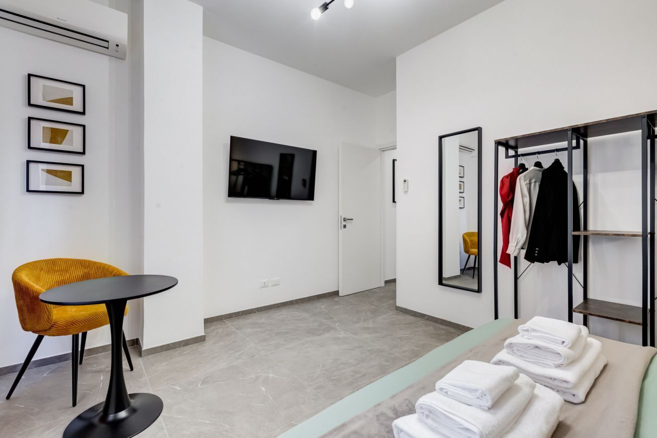 A modern hotel room with a flat-screen TV, a yellow chair, a table, a mirror, and a clothing rack with towels.