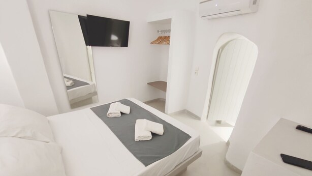 A hotel room with a bed, a flat-screen TV mounted on the wall, a towel on the bed, a small table with a remote control, and a white wardrobe. - Nolis White Fira (Santorini)
