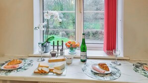 Une table à manger dressée avec de la nourriture et des boissons, une fenêtre donnant sur un paysage verdoyant et un rideau rouge.