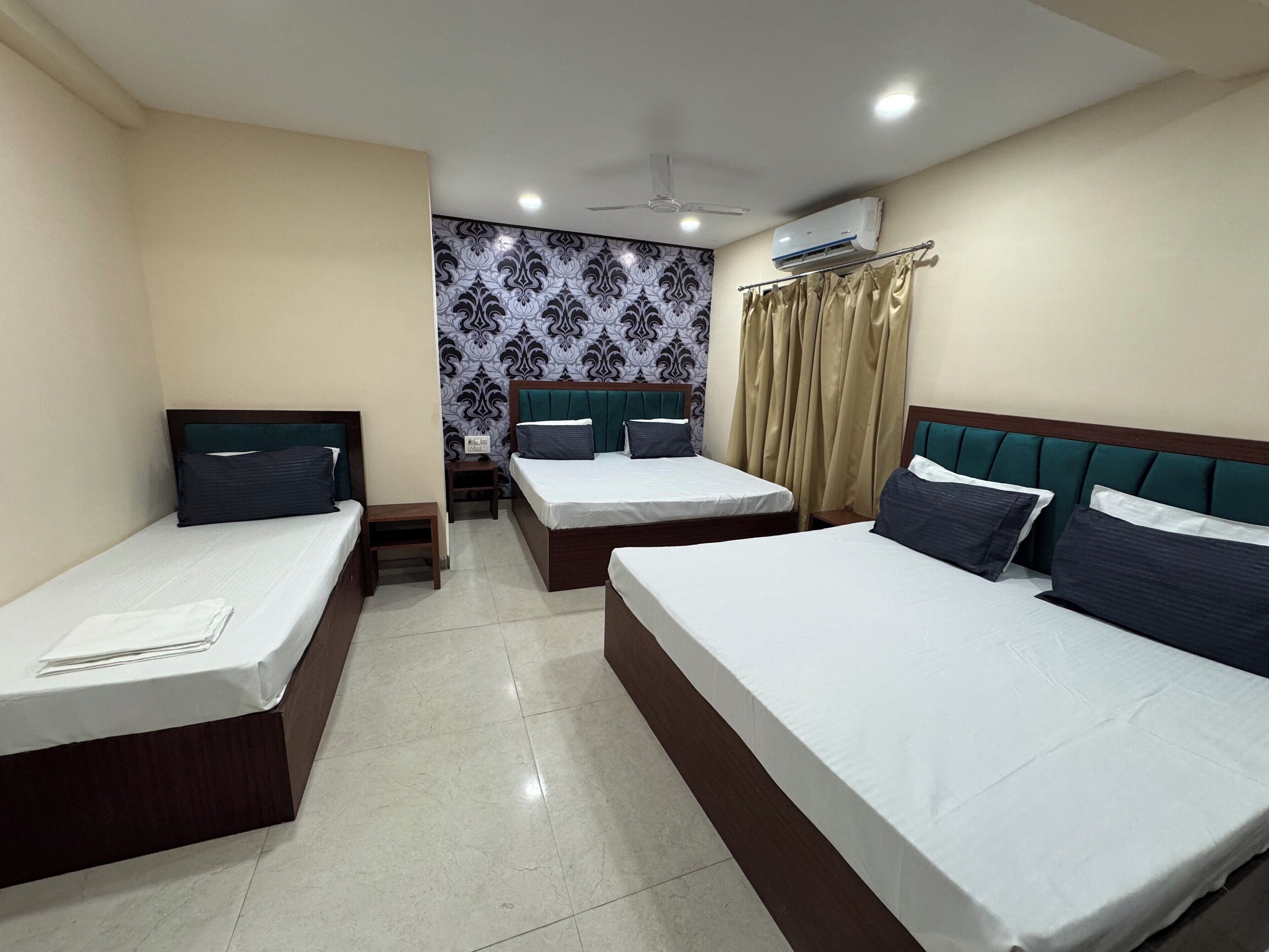 Habitación de hotel con tres camas individuales, una mesita, una mesita de noche, un ventilador de techo y un equipo de aire acondicionado.