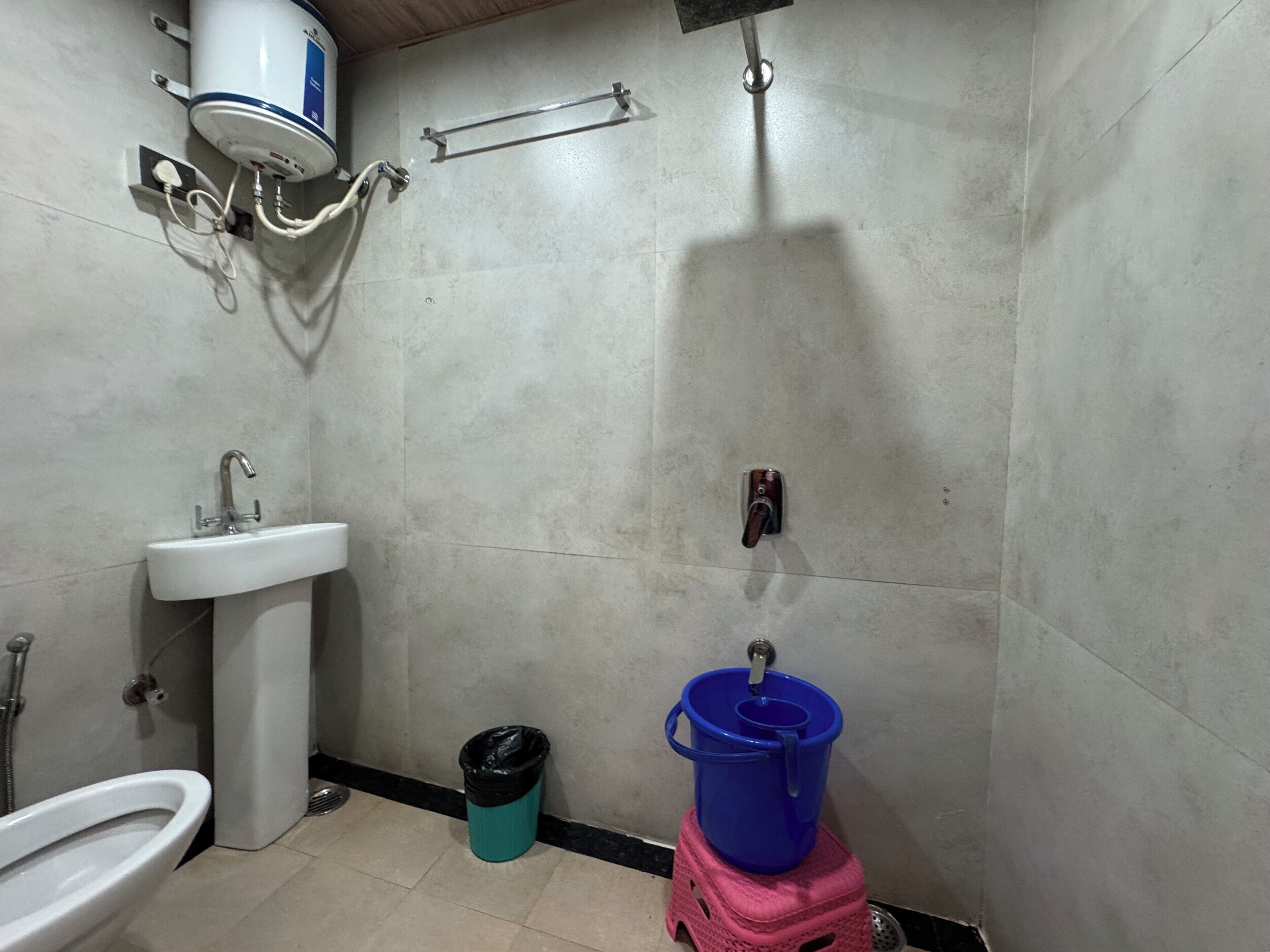Un baño con lavamanos, inodoro, ducha y un cubo azul sobre un taburete.