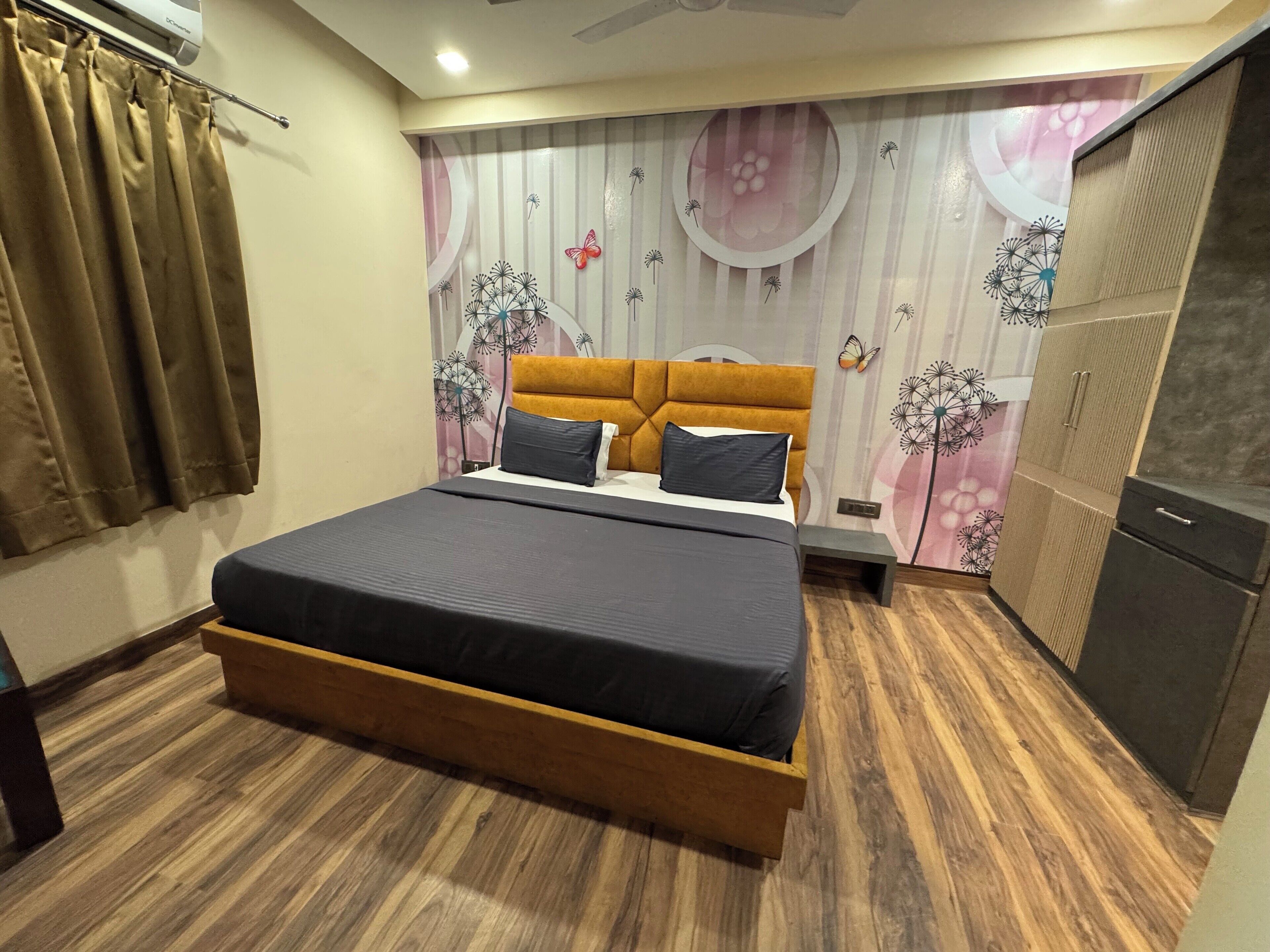 A bedroom with a bed, wooden floor, curtains, a wardrobe, and a wall with floral and dandelion patterns.
