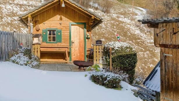 En træhytte med grøn dør og vindueskasser, en træterrasse, og et sneklædt landskab.