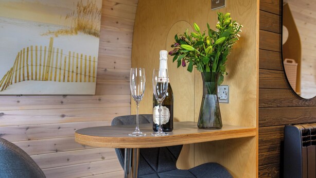 A wooden table with two wine glasses and a bottle, a vase with flowers, and two grey chairs in a room with wooden walls and a painting.