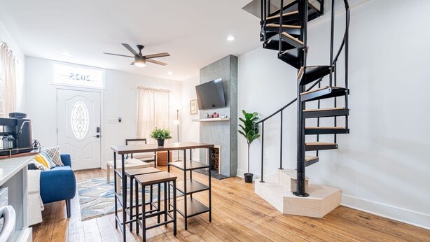 A modern living space with a wooden floor, a kitchen area, a dining table, a blue sofa, a staircase, and a ceiling fan.