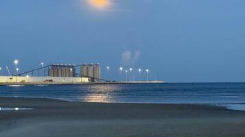 Een strand met een grote industriële structuur op de achtergrond, een waterpartij en een heldere lucht.