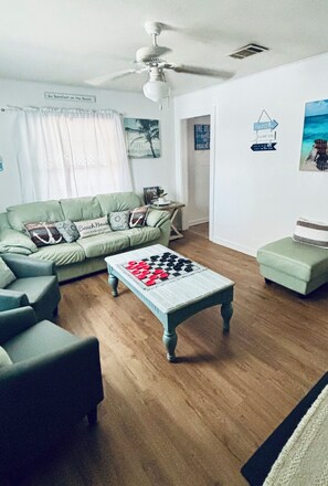A living room with a green sofa, a coffee table with a checkered pattern, a ceiling fan, and a sign on the wall.