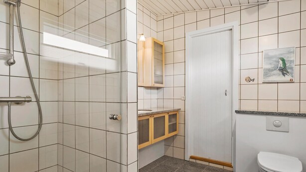 A bathroom with white tiled walls, a shower with a handheld hose, a wooden cabinet with a glass door, a white toilet, and a framed picture on the wall. - 2 bedroom gorgeous home in Juelsminde (Juelsminde)