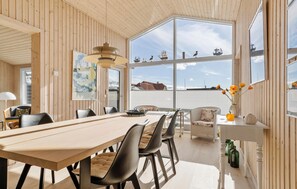 Uma sala de jantar com mesa e cadeiras de madeira, grandes janelas com vista para navios e um lustre.