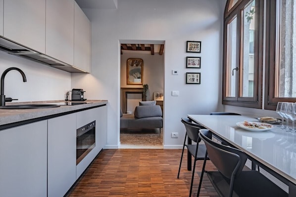 Une cuisine moderne dotée d’un four encastré, un coin repas avec une table et des chaises blanches, et une vue sur l’espace de vie, accessible par une porte ouverte, où se trouvent un canapé et un tableau.