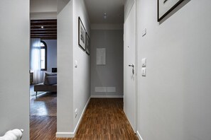Un couloir doté d’un plancher en bois, de murs blancs et d’une porte donnant sur une pièce où se trouve un canapé gris.